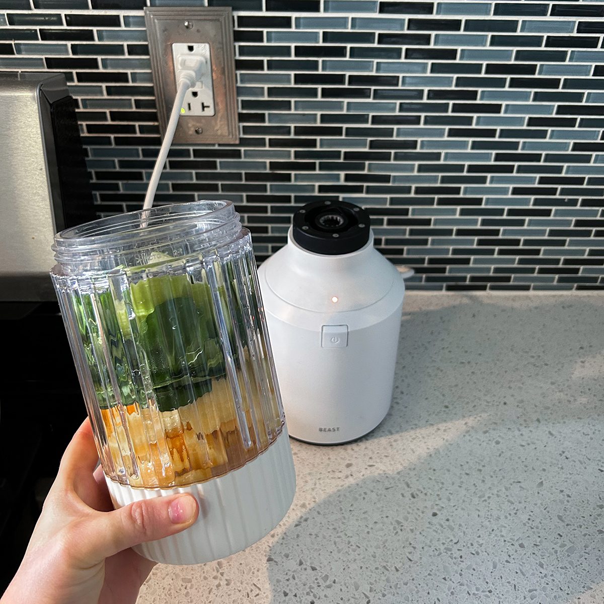 A hand holding a blender cup filled with spinach leaves, cucumber, and pineapple. The base of the blender is on the countertop in front of a tiled backsplash. An electrical outlet is visible above the countertop.