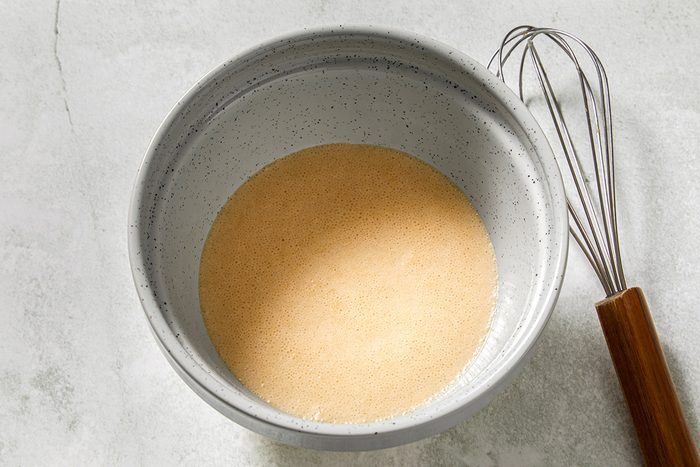 A speckled mixing bowl filled with a tan batter sits on a light gray surface. A metal whisk with a wooden handle rests beside the bowl.