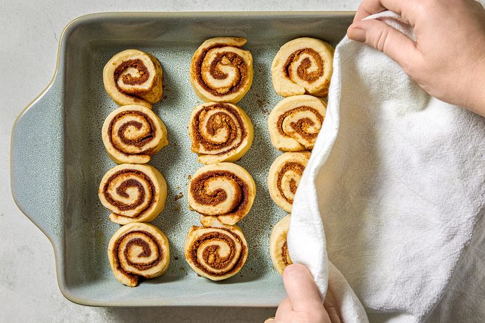 A person covers a baking tray of uncooked cinnamon rolls with a white cloth. The rolls are arranged in three rows, ready to be baked. The tray is on a light-colored countertop.