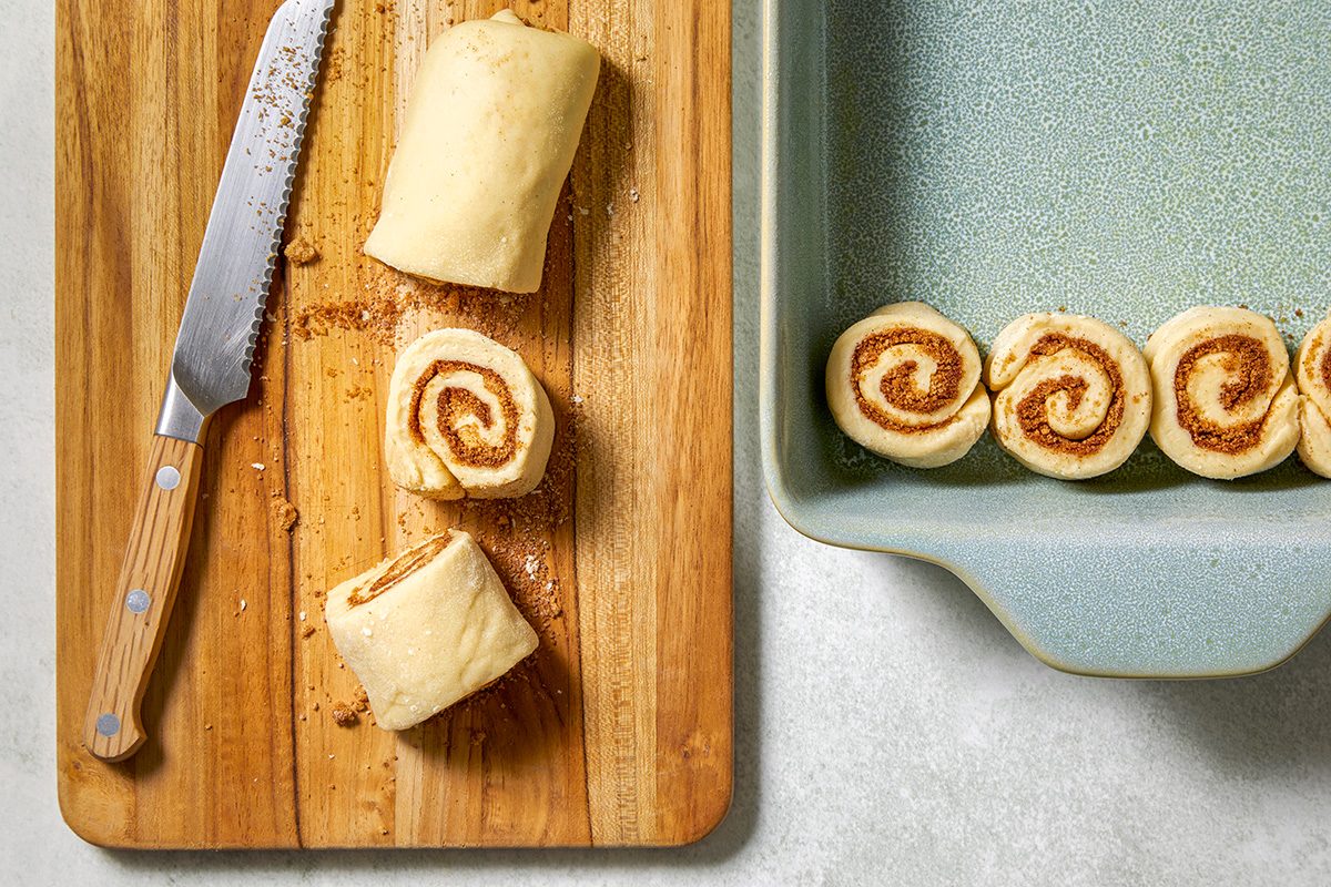 A wooden cutting board with partially sliced cinnamon roll dough next to a knife. Two rolls are visible on the board, and more are neatly arranged in a light green ceramic baking dish on the right.