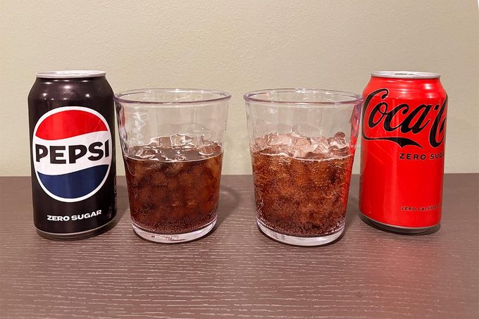 Black can of Pepsi Zero sugar with a glass of pepsi zero over ice on the left, with a Red can of Coca Cola Zero Sugar next to a glass of coca cola zero on ice on the right, both on a wooden table and beige background