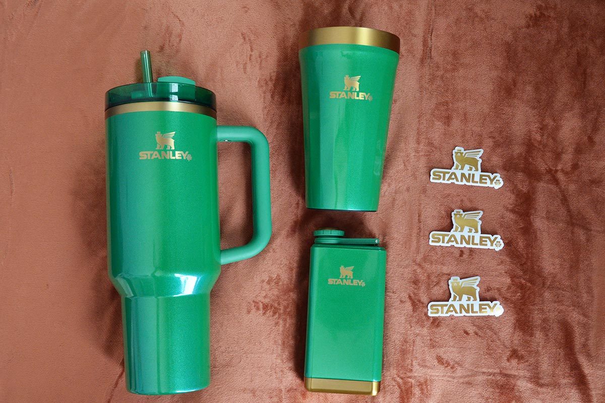 Three green Stanley drink containers with three Stanley pins on a brown fabric background