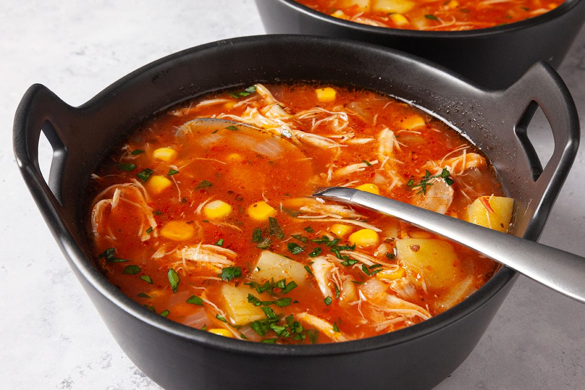 A black bowl filled with vibrant red soup containing chunks of chicken, corn, potato, and herbs. A spoon rests inside the bowl. The background shows a similar bowl, partially visible.
