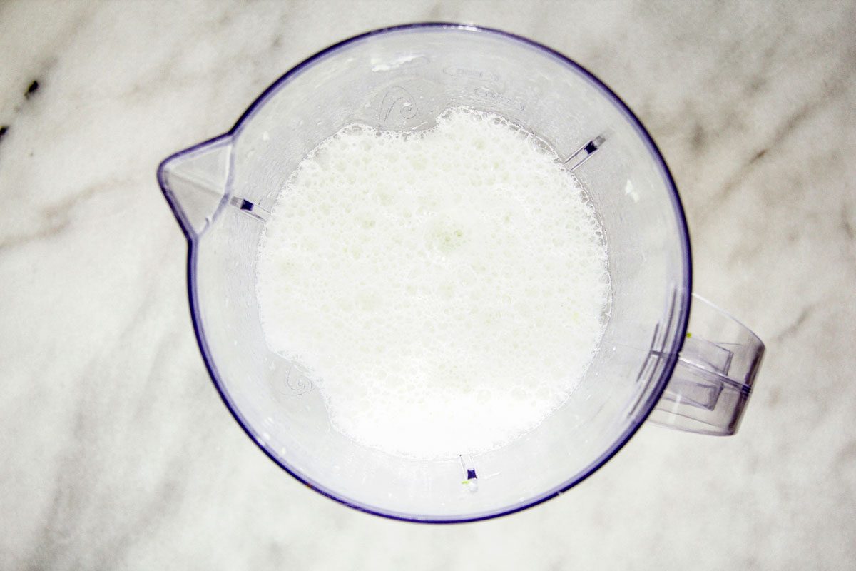A top view of a transparent blender filled with granulated white sugar. The blender is placed on a white marbled surface.