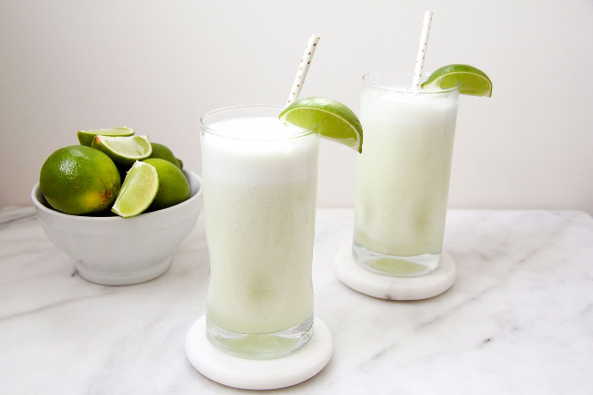 Two tall glasses of Brazilian Lemonade, garnished with lime wedges and straws, sit on round white coasters on a marble surface.