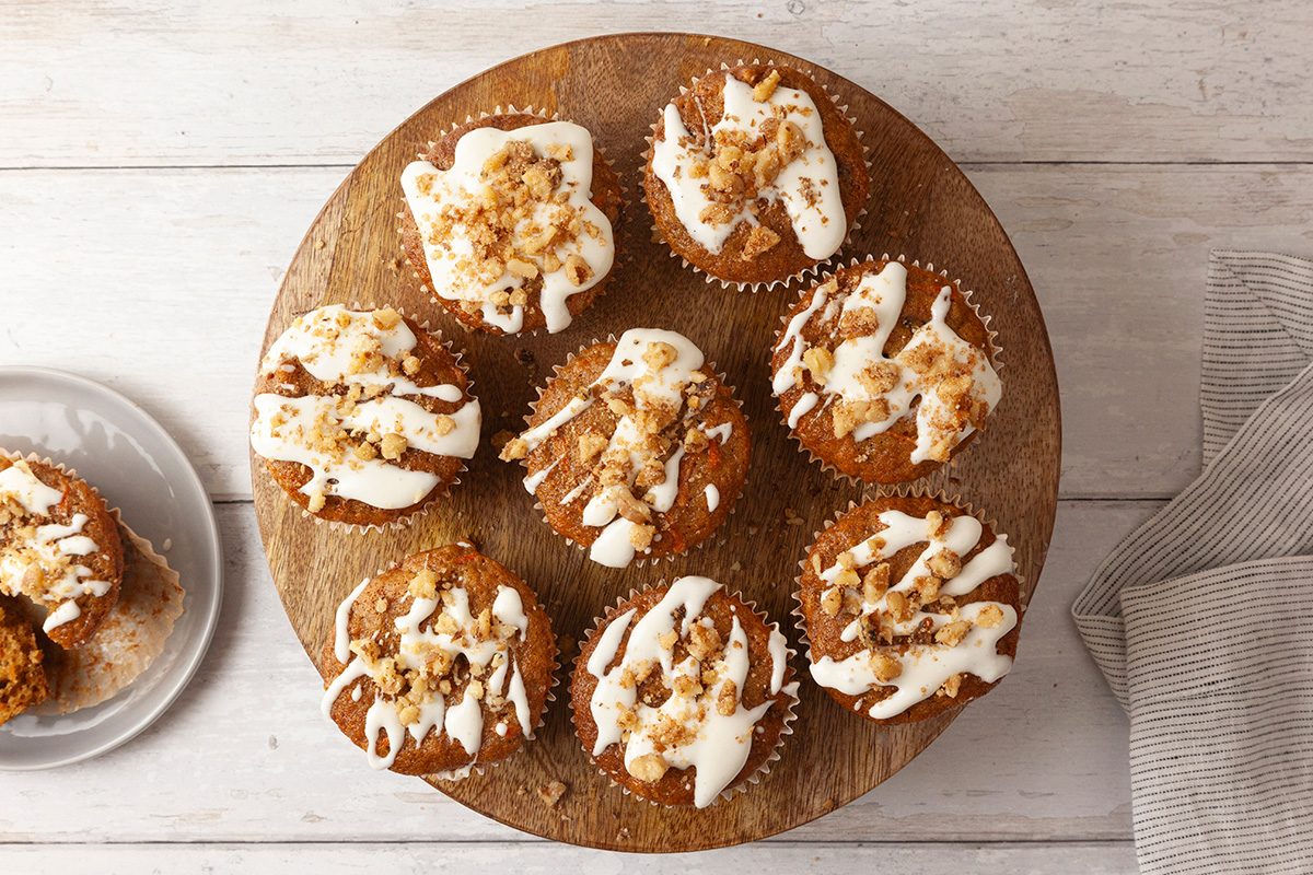Seven muffins with a white glaze drizzle and nut toppings are arranged on a round wooden board. One muffin is partially eaten on a white plate nearby. A striped cloth is visible on the right. The background is a white wooden surface.