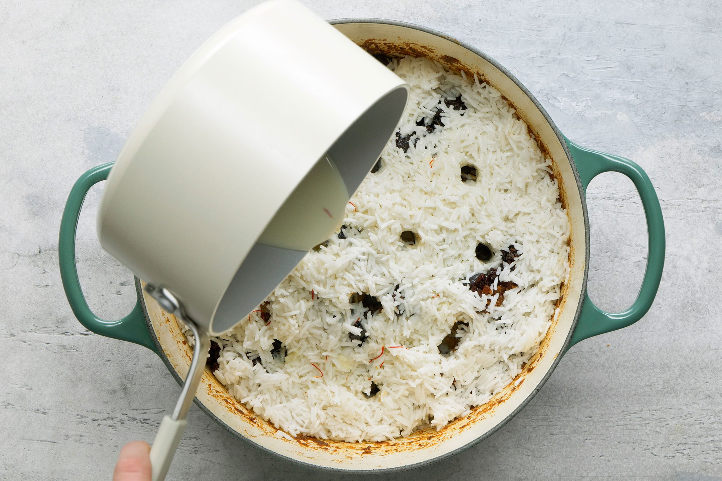 Pouring warm saffron milk over layers of rice and chicken in a dutch oven