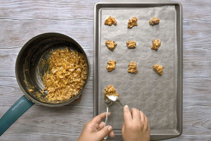 overhead shot of a person preparing a batch of homemade caramel candy; a pan with the caramel and nuts mixture sits on the counter next to a baking sheet lined with parchment paper; the person is scooping the gooey caramel and nuts mixture onto the baking sheet to set and cool; the baking sheet already has a few scoops of caramel and nuts mixture; the person's hand is holding a spoon with the caramel mixture