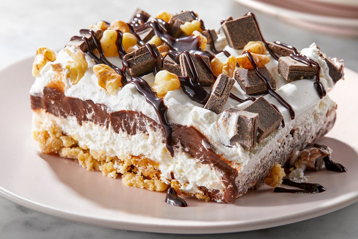 A decadent dessert bar on a plate, featuring layers of chocolate and cream filling, topped with chopped nuts, chocolate pieces, and a drizzle of chocolate syrup. 
