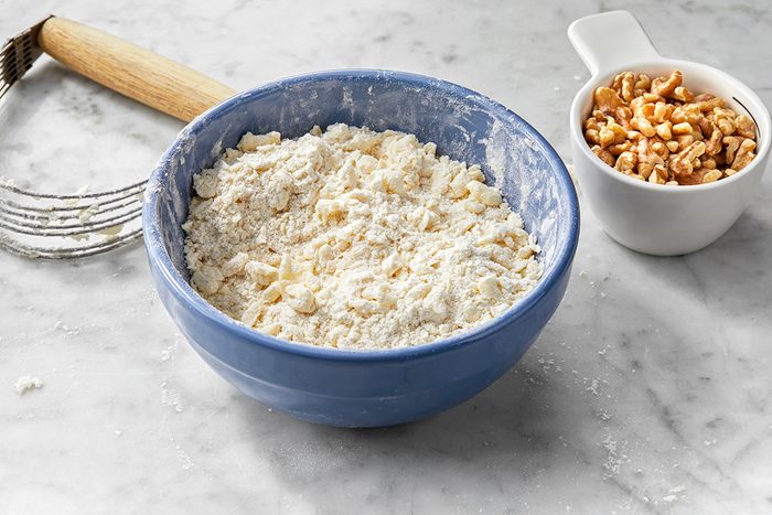 A blue bowl filled with a flour and butter mixture on a marble countertop. Beside it, a small white bowl holds chopped walnuts. A pastry cutter with a wooden handle lies nearby.