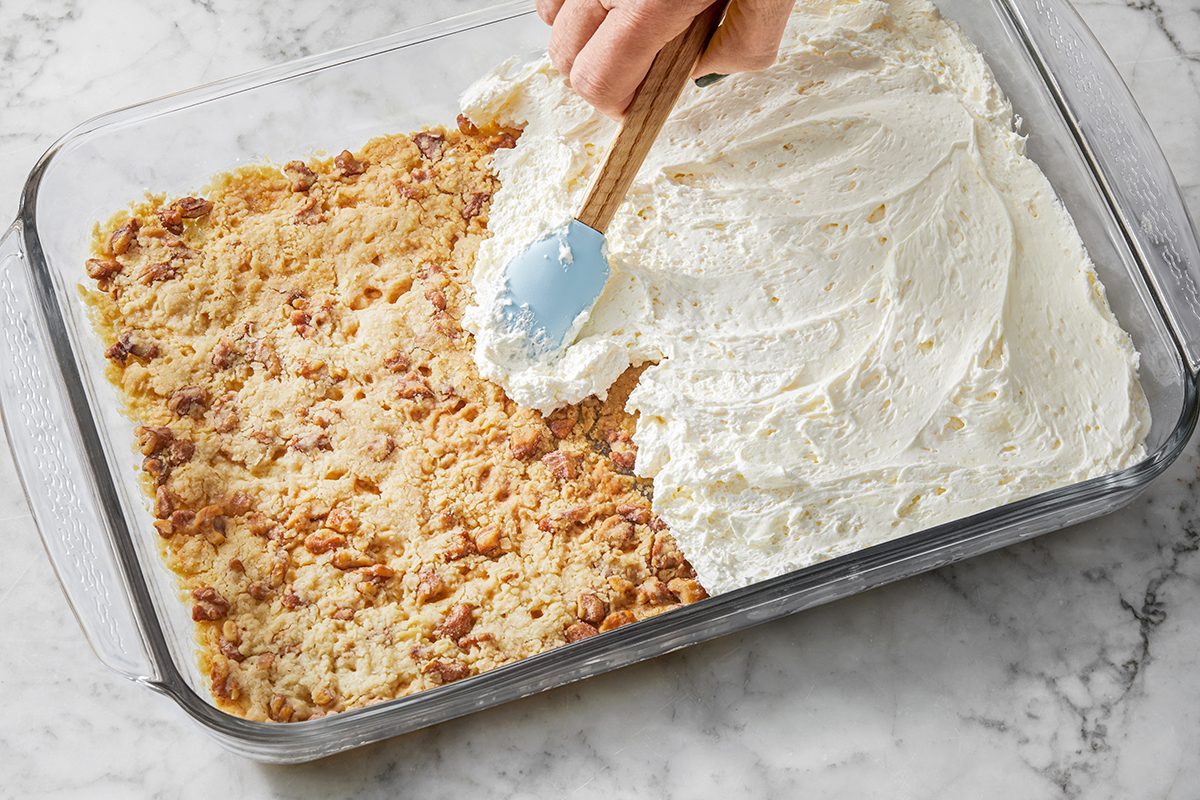 A person spreads whipped cream over a crumbly dessert topped with nuts in a glass baking dish, on a marble surface.
