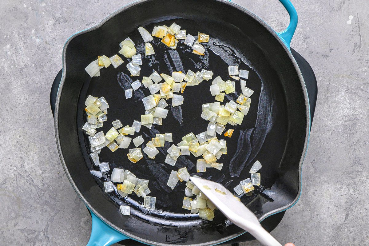 overhead shot of diced onions and minced garlic being sauteed in a black pan, a white spatula is being used to stir the vegetables, the pan is set on a textured grey surface