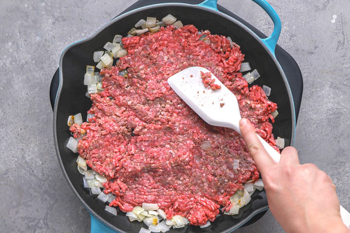overhead shot of ground beef being added to a black pan, the pan already contains sauteed diced onions and minced garlic, and a white spatula is in the process of breaking up and mixing the raw ground beef with the vegetables, the pan rests on a textured grey surface