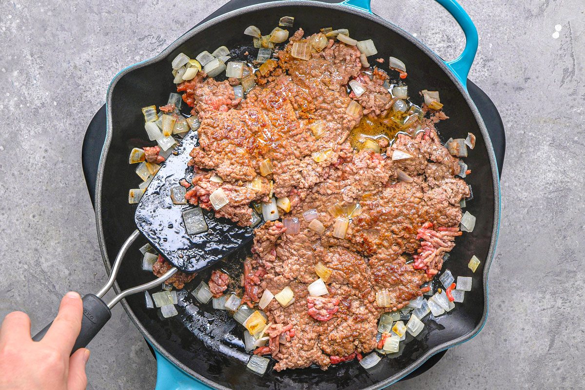 overhead shot of ground beef being cooked in a black pan with a light blue handle alongside diced onions and minced garlic, a black spatula is being used to stir and break apart the ground beef, the pan sits on a textured grey surface,