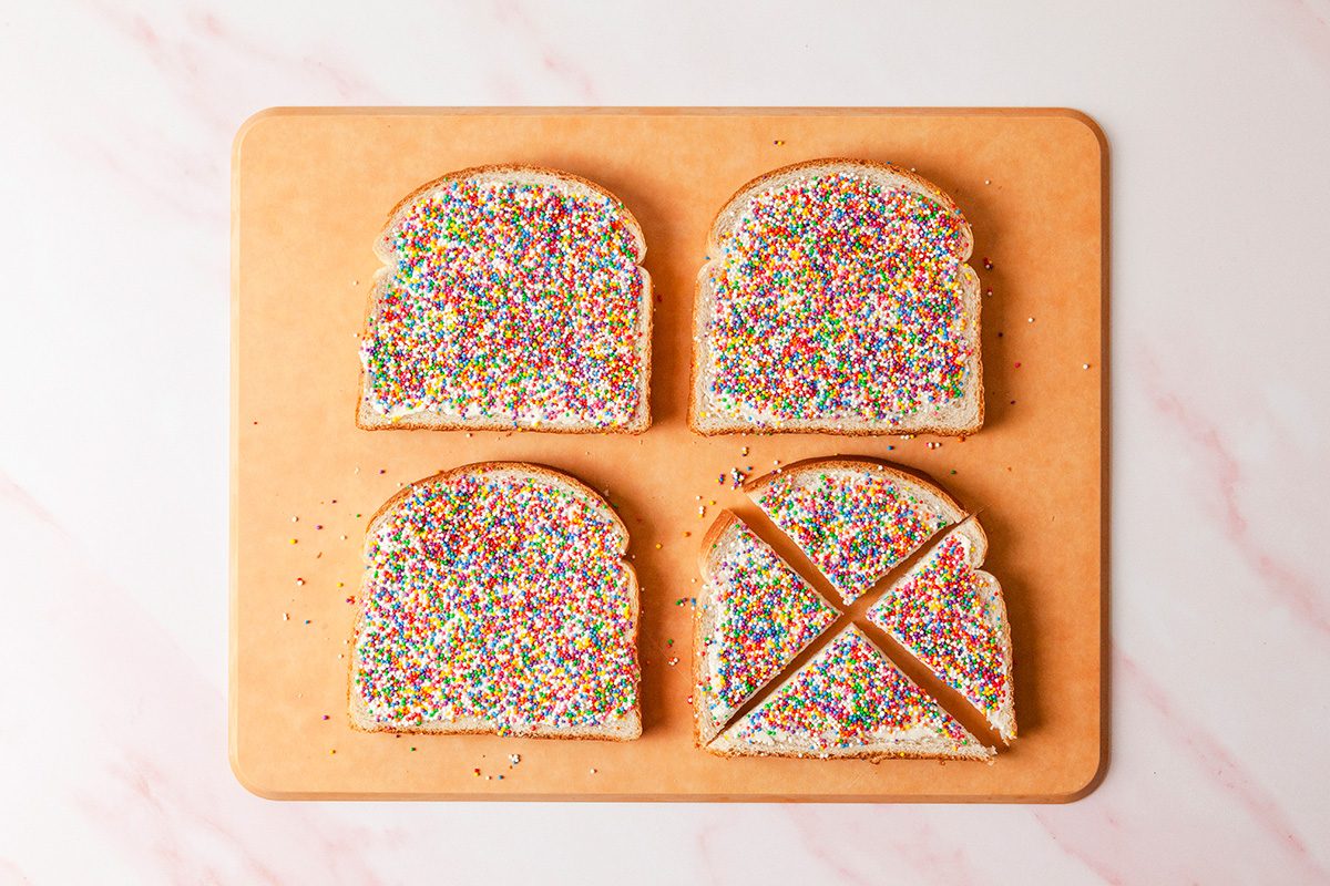 Four slices of bread covered with colorful sprinkles are placed on a wooden cutting board. Three slices are whole, while one is cut into two triangles. The background is a light, marbled surface.