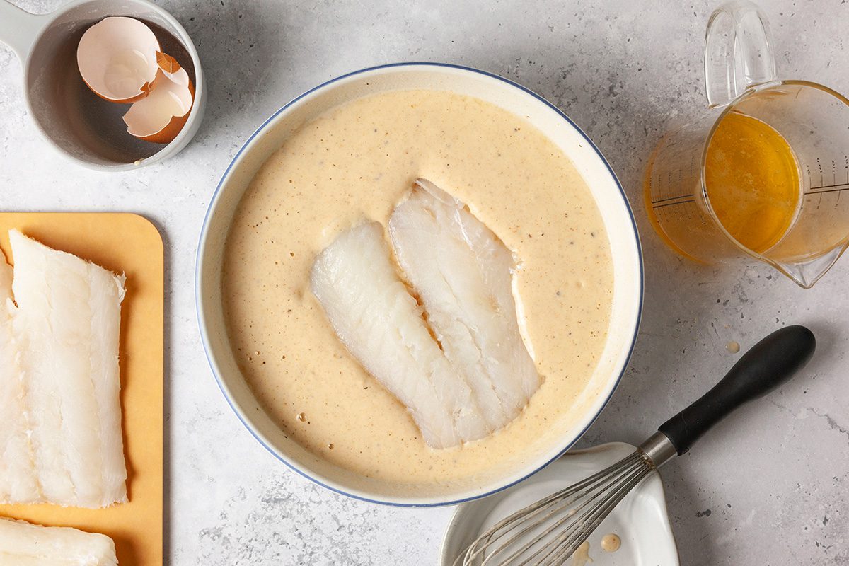 A bowl of beige batter with two fish fillets inside. To the left, more fish fillets on a cutting board. A cup with eggshells and a measuring jug with orange liquid are nearby. A whisk and bowl are at the bottom right on a light countertop.