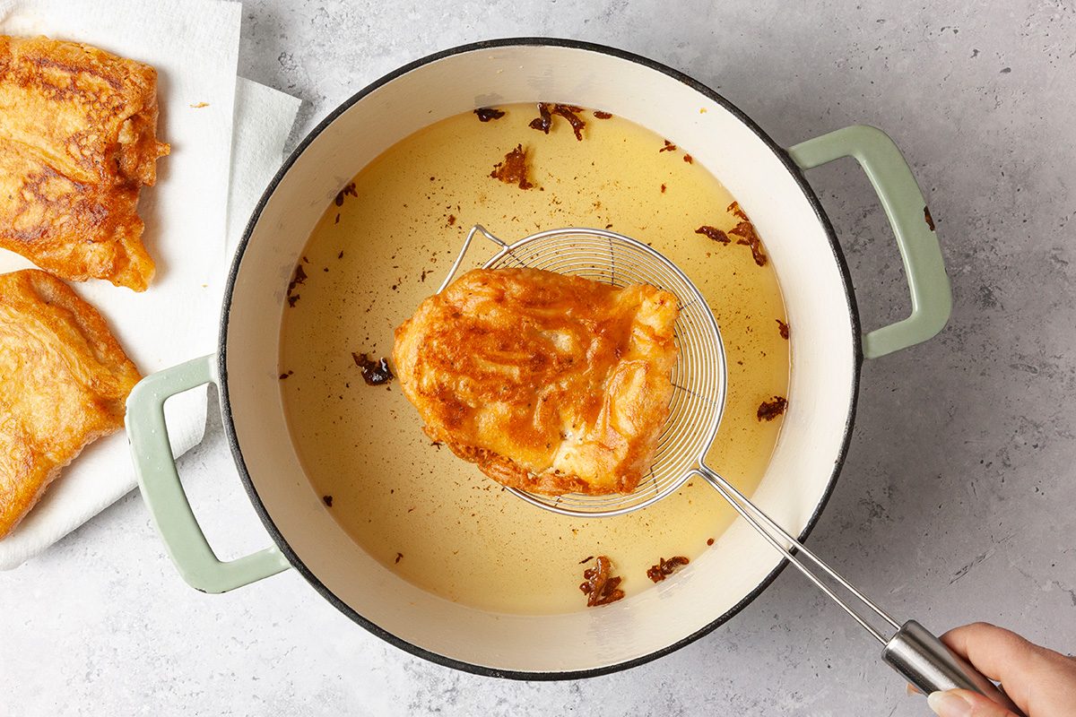 A piece of fried chicken is being lifted from a pot of oil using a slotted spoon. The pot is light green with handles. There are other pieces of chicken resting on a paper towel nearby. The surface is a light gray.