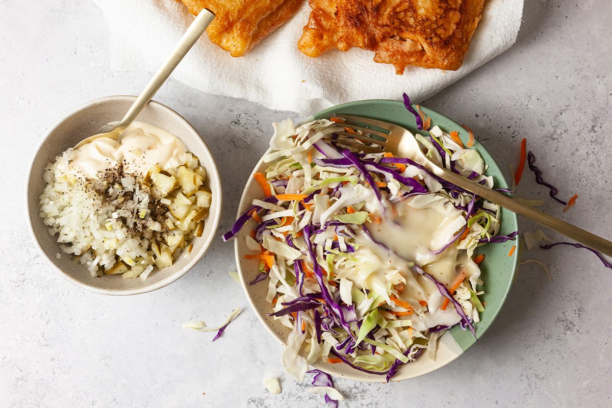 A bowl of coleslaw with purple cabbage, carrots, and creamy dressing next to a small bowl of diced potatoes and spices. Fried food is partially visible on a paper towel in the background.
