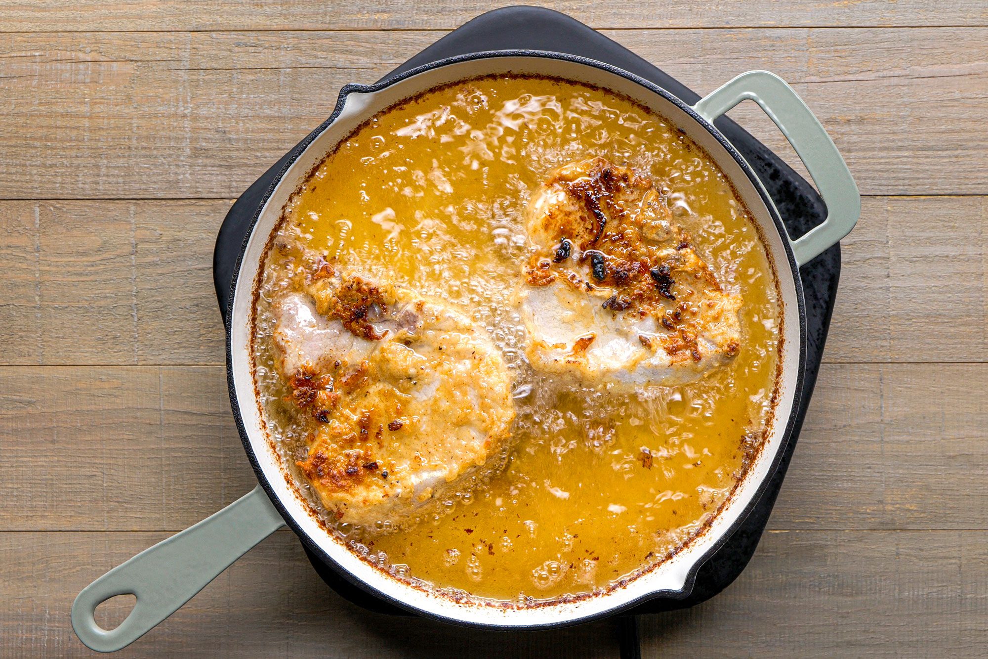 overhead shot of a cast iron skillet filled with sizzling oil, in which two pieces of breaded meat are being fried; the skillet is placed on a wooden surface