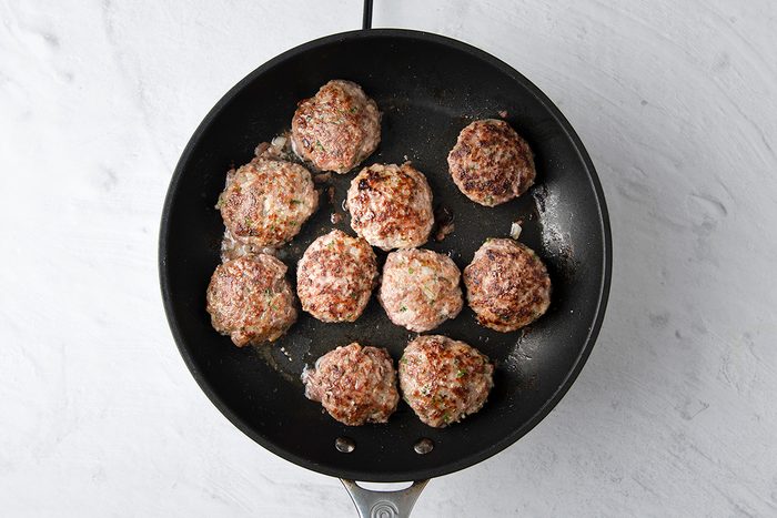A non-stick skillet with nine cooked round meat patties, slightly browned, arranged on a light gray countertop.