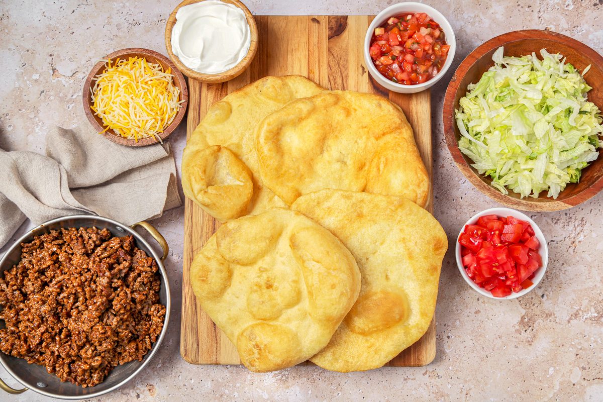 Frying bread ready to be topped with meat mixture, lettuce, and tomato, served with salsa and sour cream