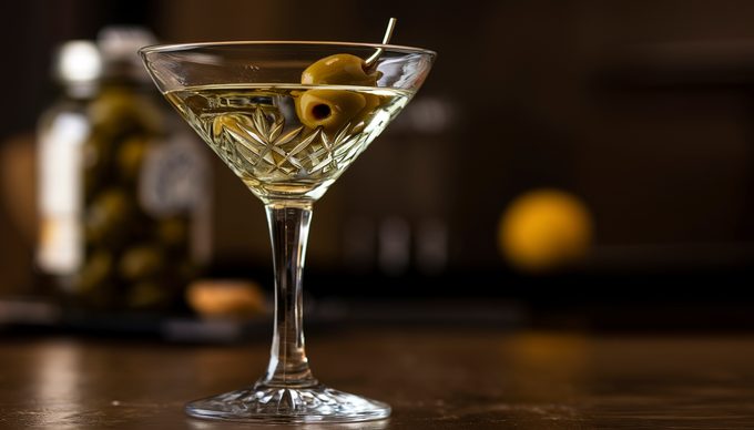 Yellow-tinged martini in a crystal glass with an olive, sitting on a dark bar