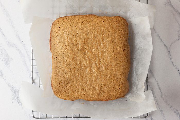 A square gingerbread sheet cake cooling on a wire rack, lined with parchment paper. The cake has a golden-brown color and sits on a light marble surface.