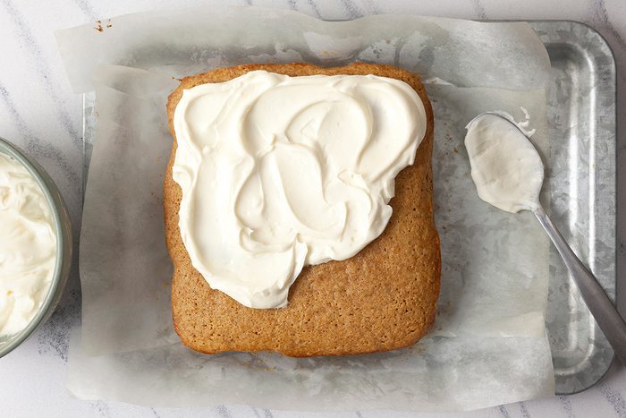 A square cake on a sheet of parchment paper partially frosted with white icing. A spoon with icing rests beside the cake on a metal tray. A bowl of icing is partially visible in the corner.