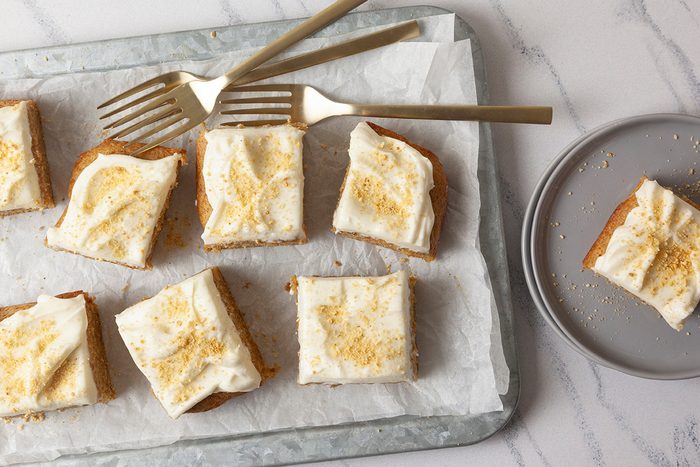 Squares of frosted cake topped with a sprinkle of crumbs are arranged on a parchment-lined tray. Three forks are placed beside the cakes.