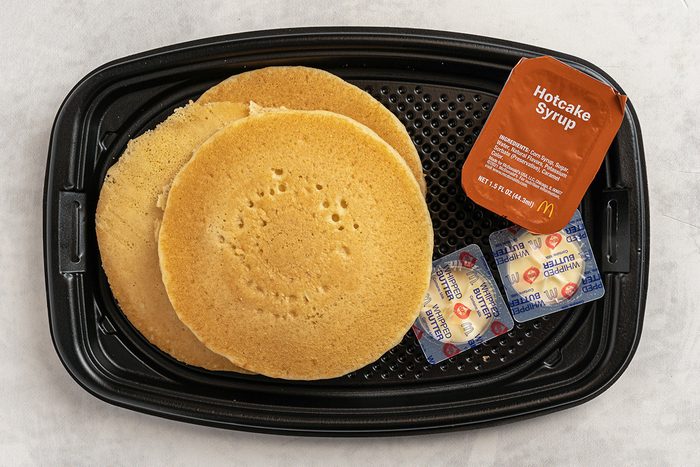 A black tray with three golden-brown pancakes, a container of hotcake syrup, and two butter packets on a light surface.
