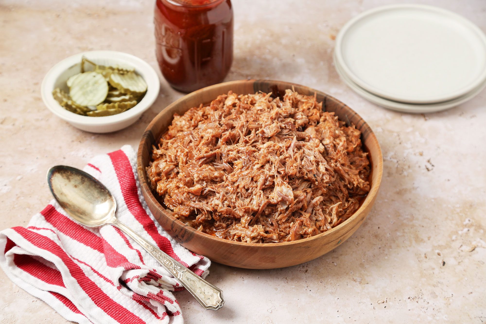 3/4 angle view shot of Instant Pot Pulled Pork; served in a large wooden bowl; with a napkin and a spoon; two empty white plates and barbeque sauce are nearby; all set on a marble surface;
