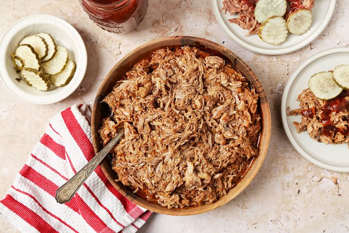 Overhead shot of Instant Pot Pulled Pork; In a large wooden bowl wth spoon; served on two white plates with barbeque sauce and pickles; a napkin nearby; all set on a marble surface;