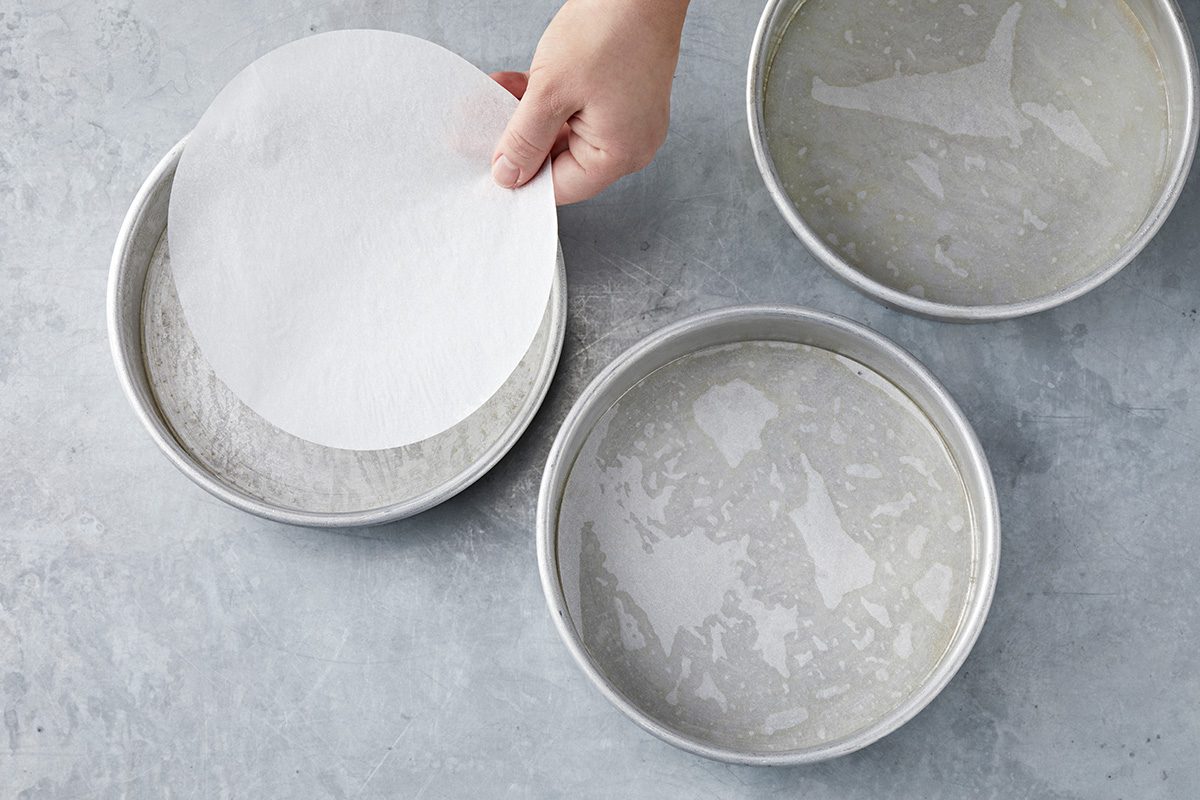 A hand places a round piece of parchment paper into one of three round cake pans. The pans are arranged on a grey surface, each appearing to be greased and prepared for baking.