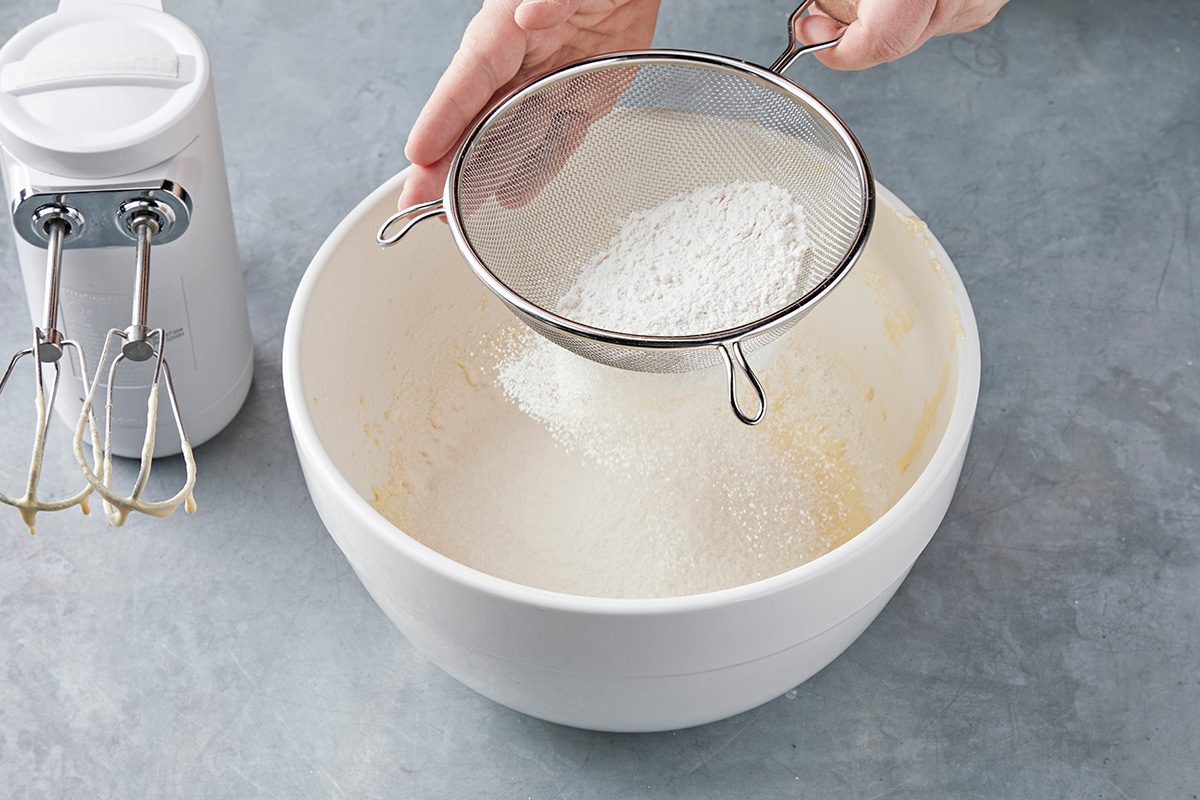 A hand holding a sifter is sprinkling flour into a large white mixing bowl on a gray countertop. An electric mixer with beaters is placed next to the bowl.