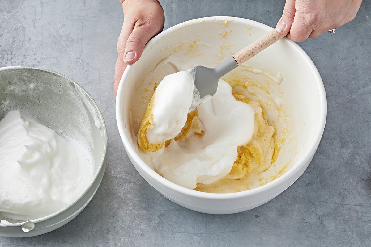 A person is using a spatula to fold beaten egg whites into a yellow mixture in a white bowl. Next to it, another bowl holds more beaten egg whites. The surface is gray and smooth.