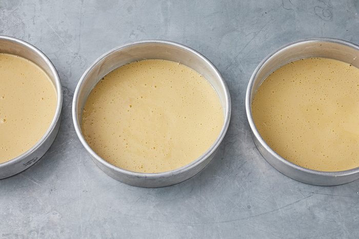 Three round cake pans filled with smooth, light yellow batter are placed side by side on a gray surface.