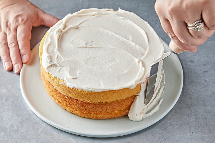 A person applies white frosting to a round yellow cake with a spatula on a white plate. The cake is on a gray surface, and the person's hands are visible, one holding the plate steady.