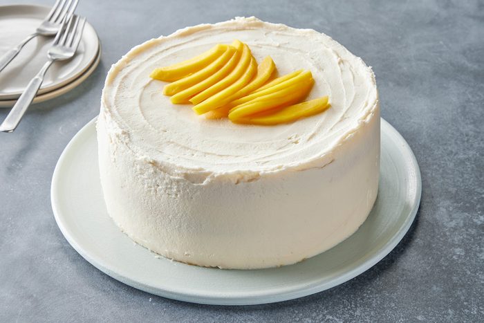 A round cake with smooth white frosting, topped with slices of fresh yellow mango, displayed on a light-colored plate. In the background, there are forks and a couple of stacked plates.