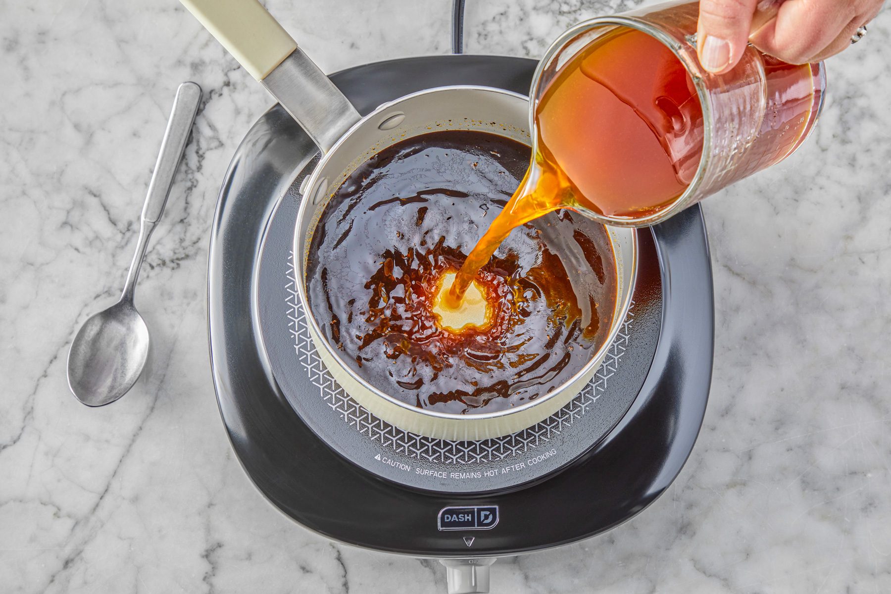 Overhead shot of pour drippings and loosened browned bits from roasting pan into a small saucepan; Skim fat; inductione stove and spoon; on a marble surface;