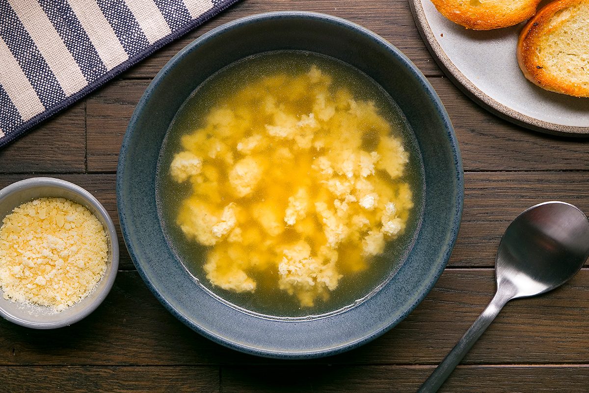A bowl of egg drop soup on a wooden table, accompanied by a small bowl of grated cheese, a spoon, and a plate with toasted bread slices. A striped cloth napkin is partially visible on the side.