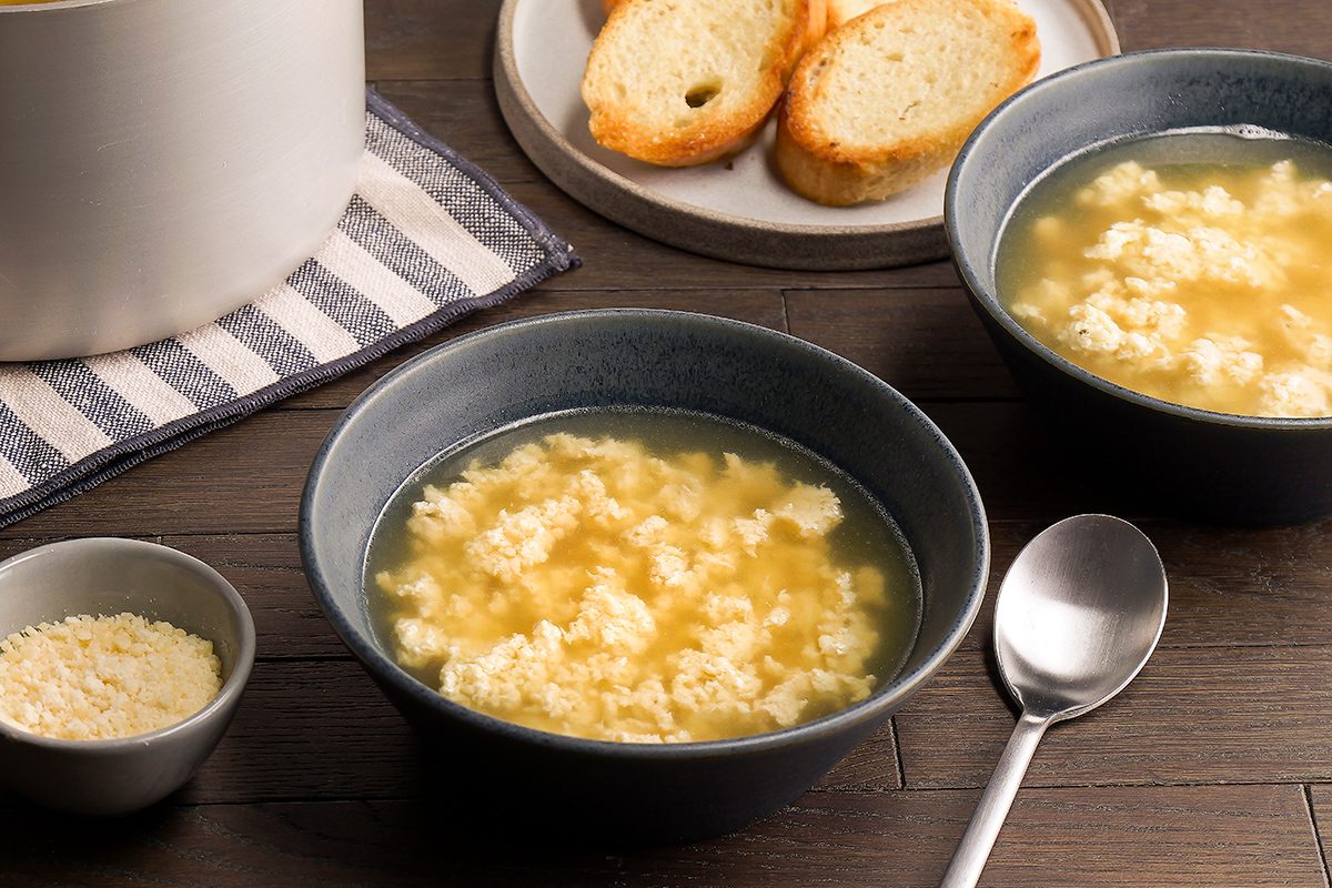 Two bowls of egg drop soup on a wooden table, accompanied by a spoon. A small bowl of grated cheese is nearby. In the background, slices of toasted bread are on a plate, and a striped napkin is partially visible.