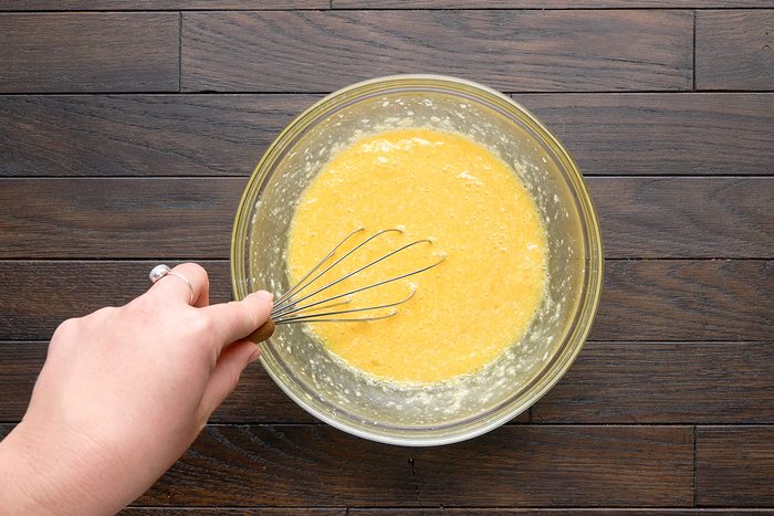 A hand whisking a yellow batter in a glass bowl on a dark wooden surface.