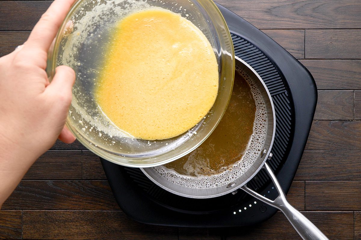 A hand pours beaten eggs from a glass bowl into a saucepan with simmering liquid on an electric stovetop. The stovetop and saucepan are on a dark wooden surface.