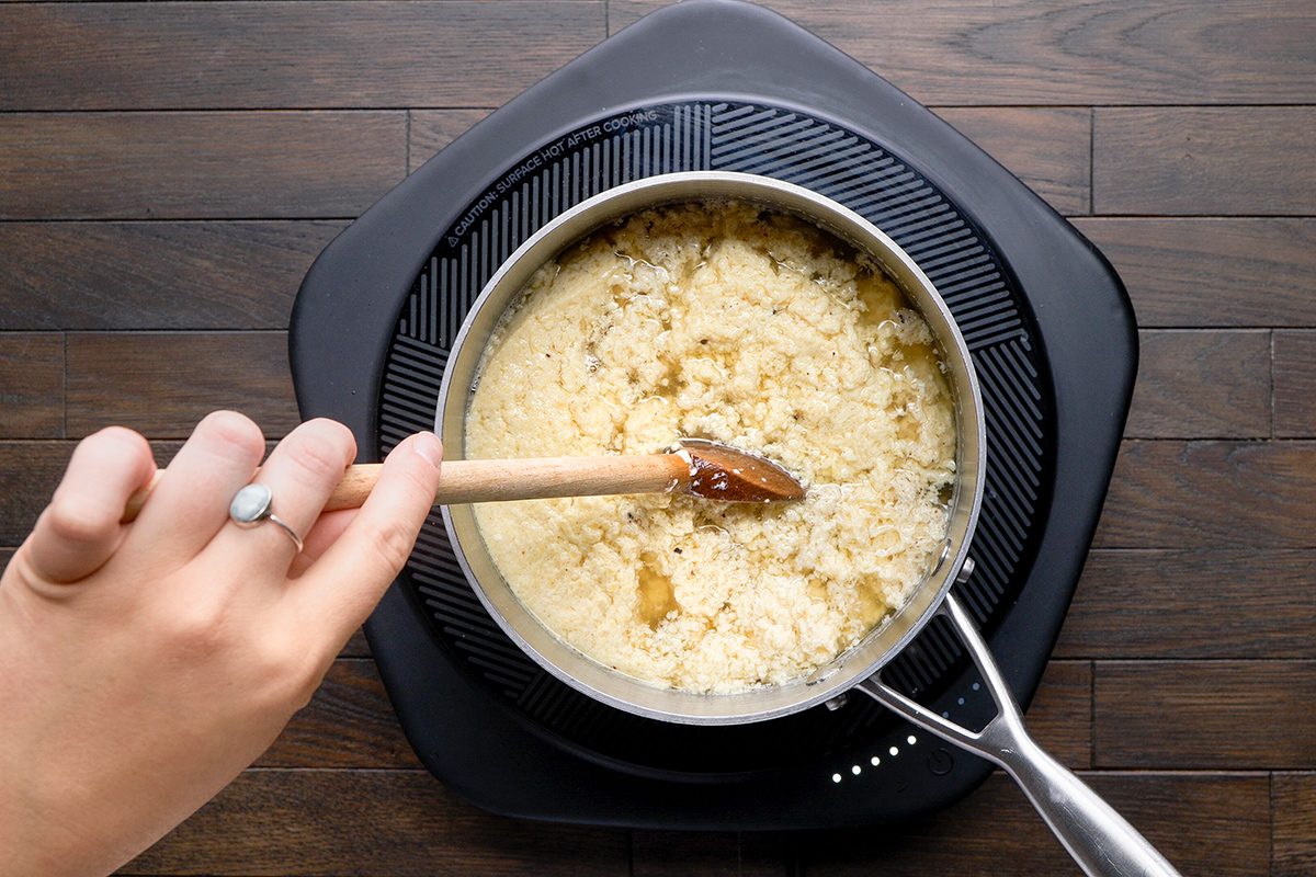 A person stirs quinoa cooking in a pot on an induction cooktop with a wooden spoon. The quinoa appears fluffy and is seasoned with visible herbs. The background features a dark wooden surface.