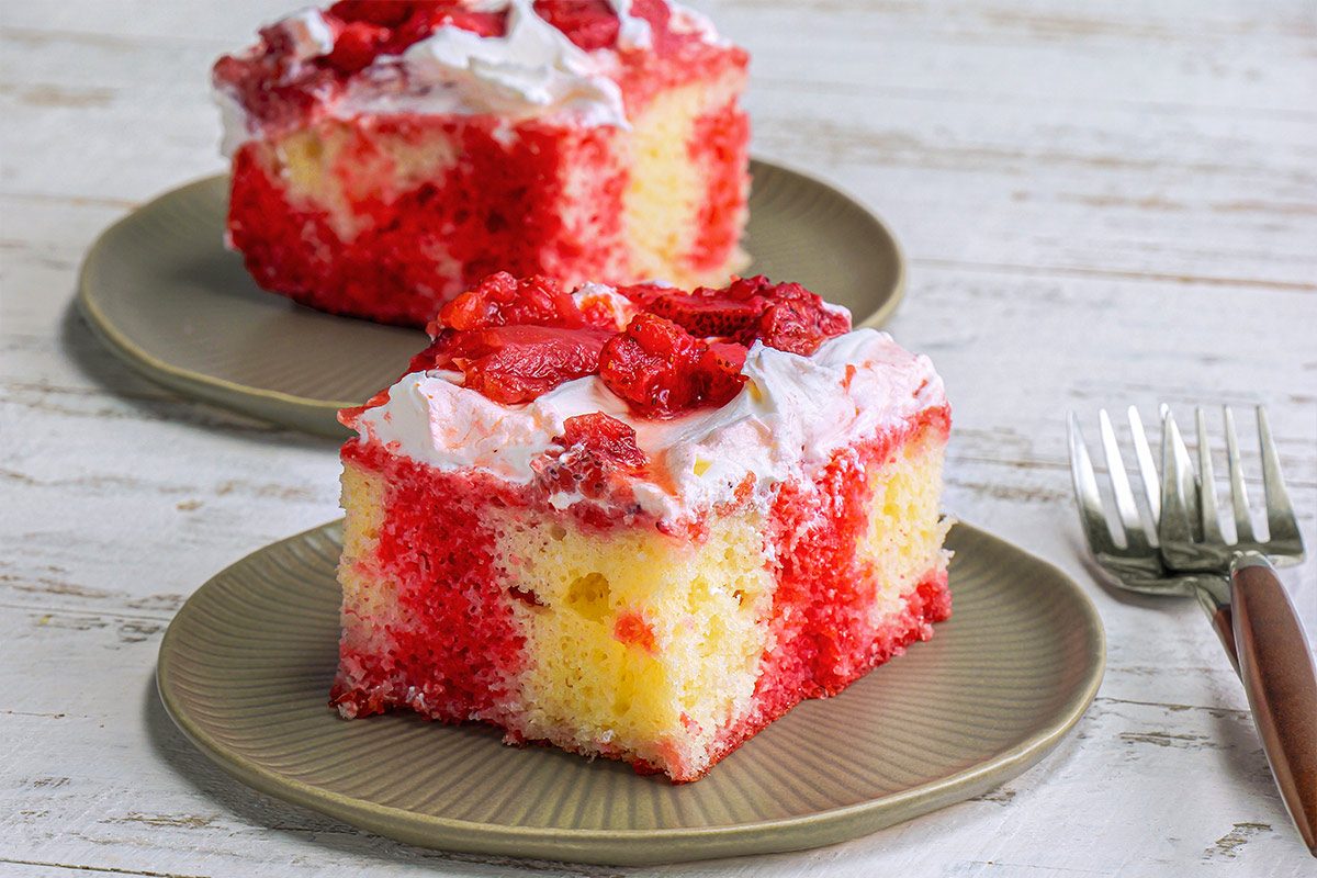 3/4th shot of cake slices served on light green plates; one slice is positioned in the foreground while the other sits slightly behind it; garnished with bright red strawberries on top; a fork with a wooden handle is placed next to one of the plates, the background is a white wooden surface