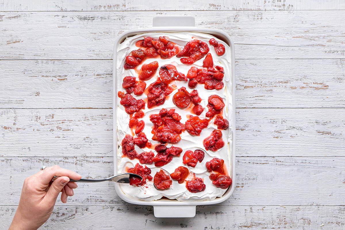 overhead shot of a rectangular dish filled with a creamy dessert layered with a topping of red strawberries and fruit syrup; a hand is seen holding a fork, the background features a rustic wooden table with a light color