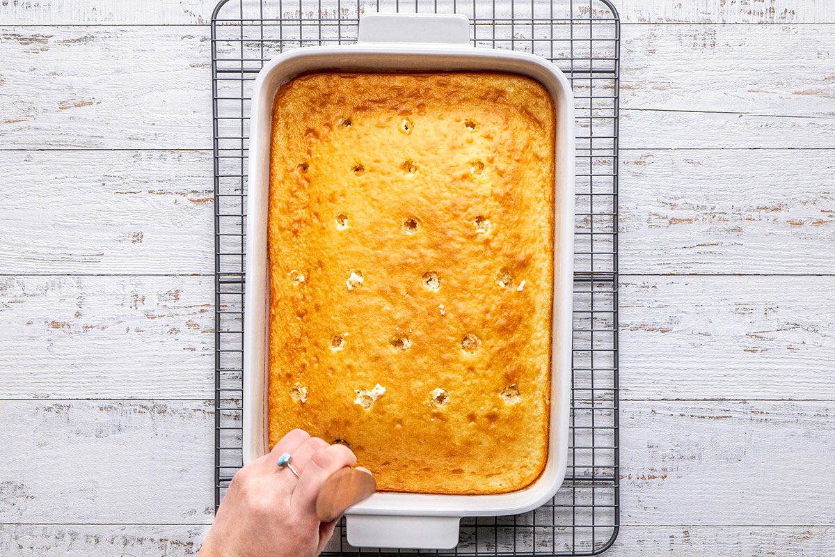 overhead shot of a freshly baked rectangular cake in a white ceramic baking dish sitting atop a wire cooling rack; a hand can be seen holding a wooden spatula, poised to cut the cake; the background is wooden tabletop