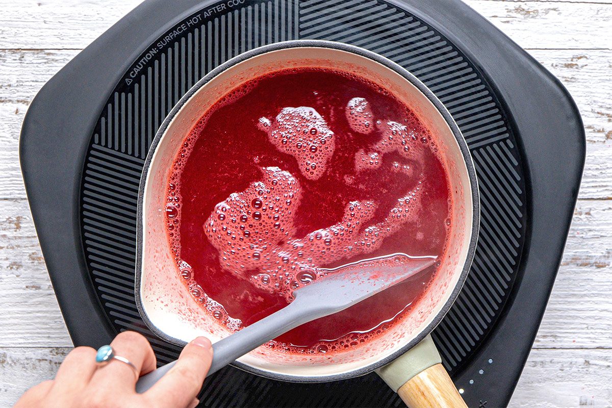 overhead shot of a pot on a heat source, filled with a rich, red liquid that is boiling and bubbling, A hand is seen holding a spatula, stirring the mixture gently with small bubbles forming on the surface; The background is a textured wooden surface