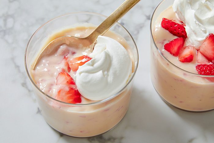 A glass of creamy yogurt topped with dollops of whipped cream and fresh strawberry slices, placed on a marble surface. A wooden spoon rests inside, partially submerged in the yogurt.