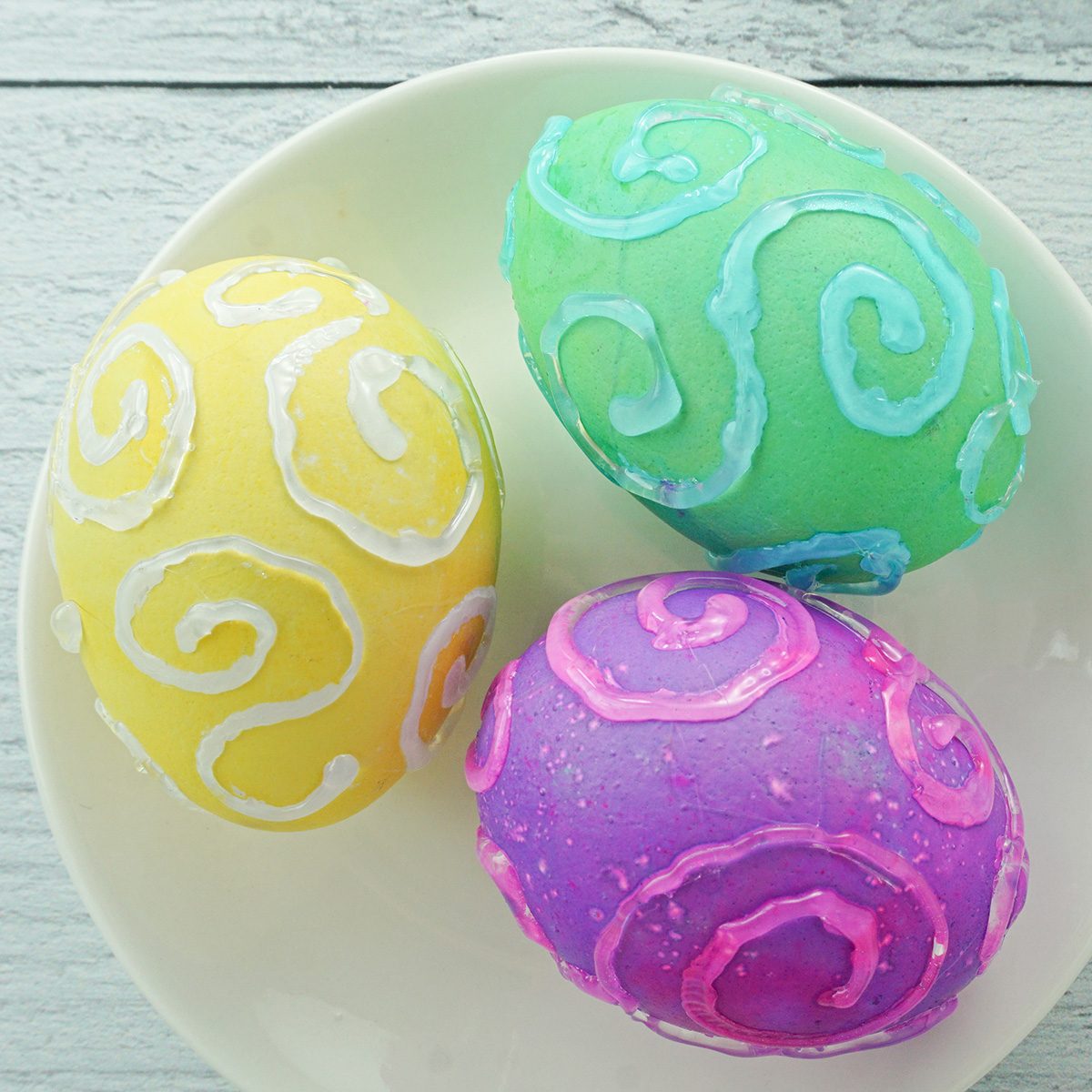 A plate with three painted Easter eggs featuring colorful swirl designs. One egg is green, another is purple with pink swirls, and the third is yellow with white swirls. They rest on a light wooden surface.